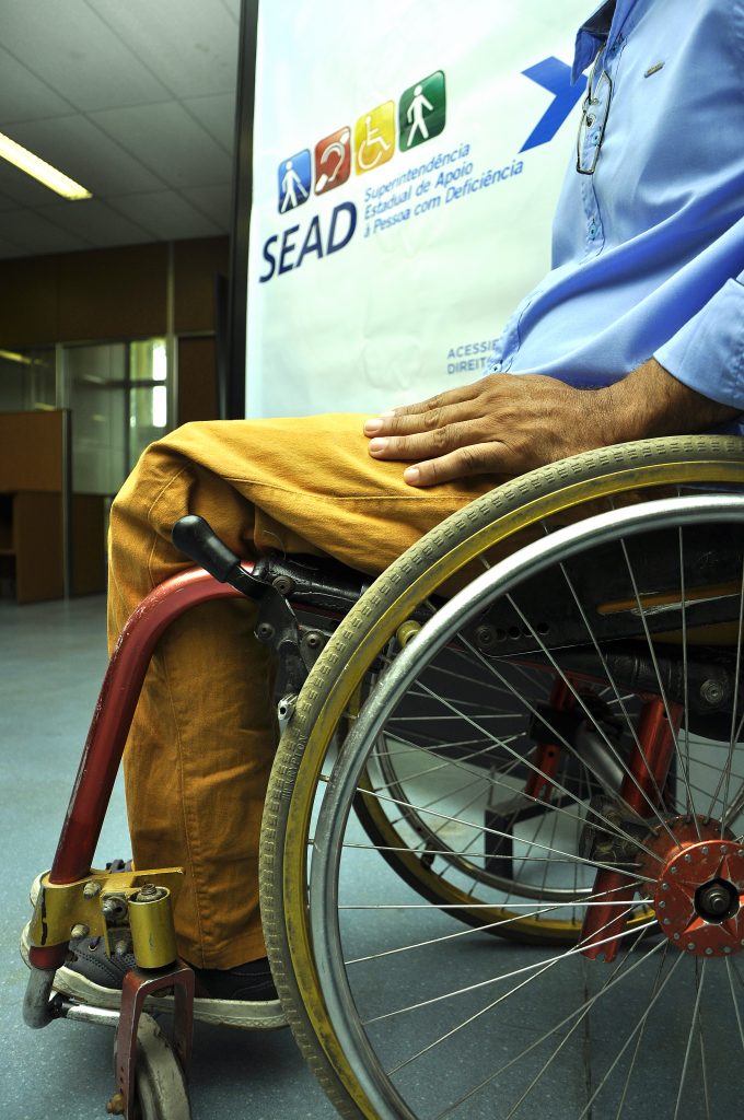 Pessoa com cadeiras de rodas em frente a um banner, que tem escrito "Sead, Superintendência Estadual da Pessoa com Deficiência"